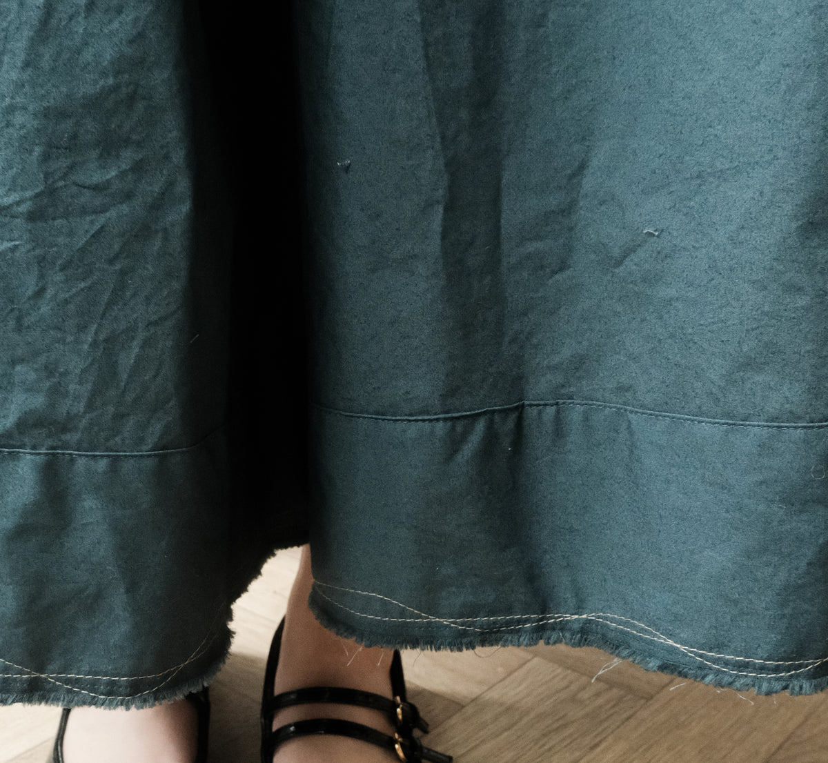 Close-up of a dark teal-green skirt with visible fabric texture, hem stitching, and a small portion of a person's feet wearing black sandals on a wood floor.