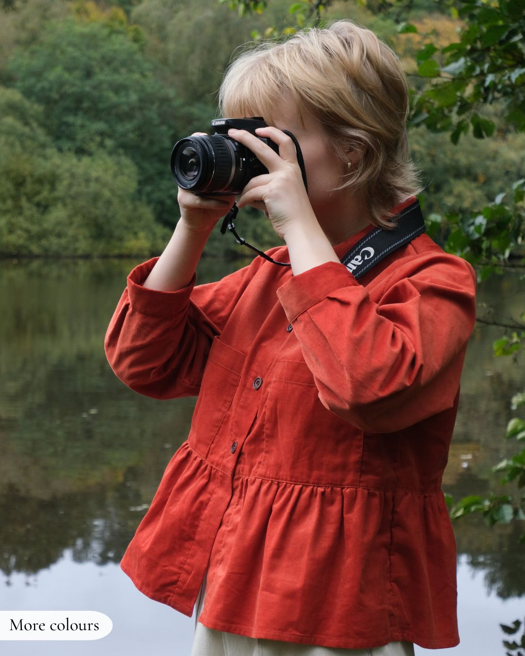 Person in a red jacket taking a photo with a camera by a body of water.