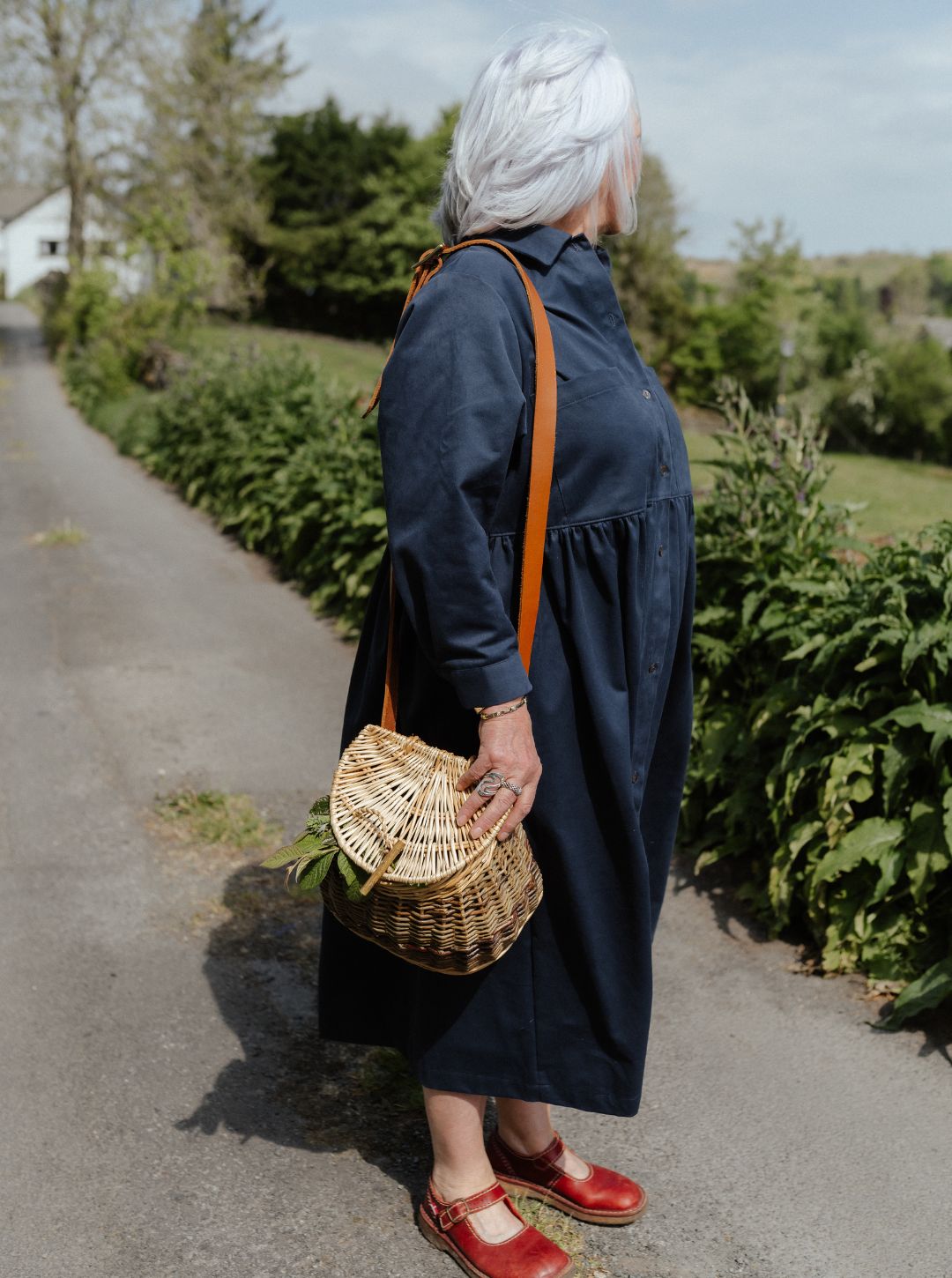 Woman in Dorothy shirtdress Noah navy blue with a straw bag walking on a path with greenery around.