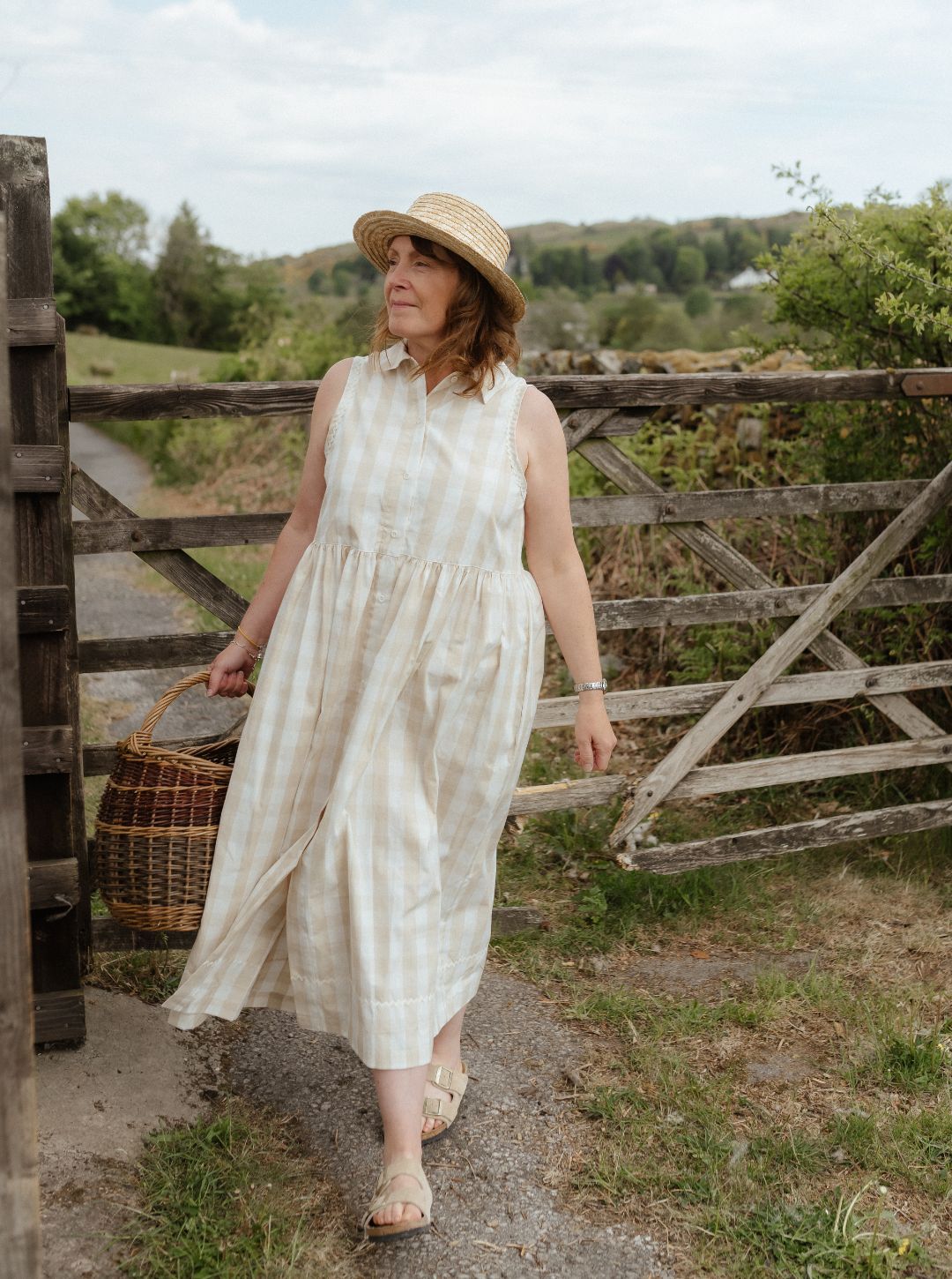 Woman in a white dress and hat standing near a wooden gate with a basket, outdoors.