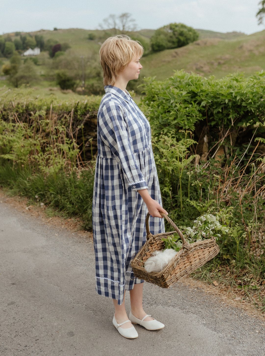 Woman in a blue checkered dress holding a basket with flowers on a rural road.