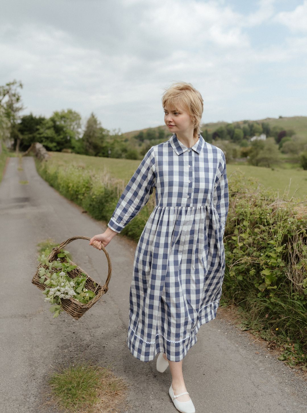 Woman in a blue checkered dress holding a basket of flowers on a rural road.