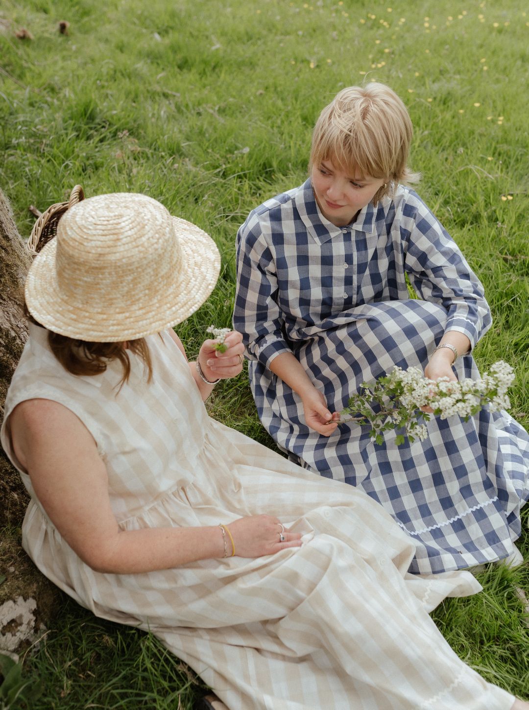 Two women sitting on grass, one in a natural check dress and straw hat, the other in a blue checkered dress, holding flowers.