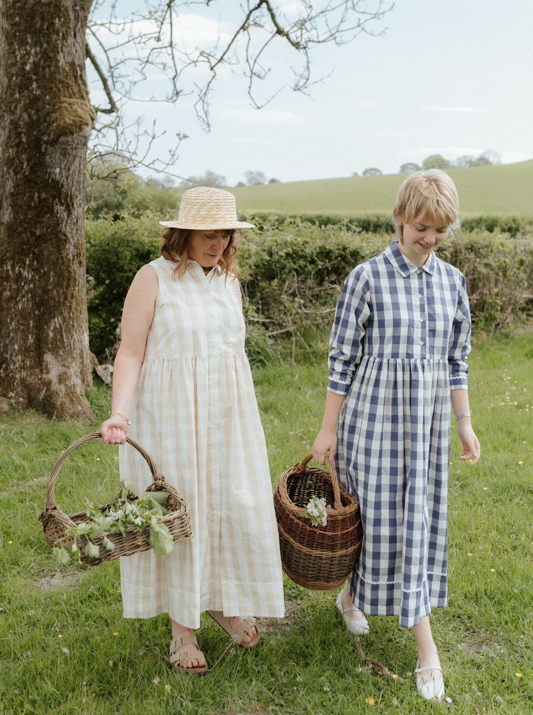 Two women in a garden holding picnic baskets, one in a natural check dress and straw hat, the other in a blue checkered dress.