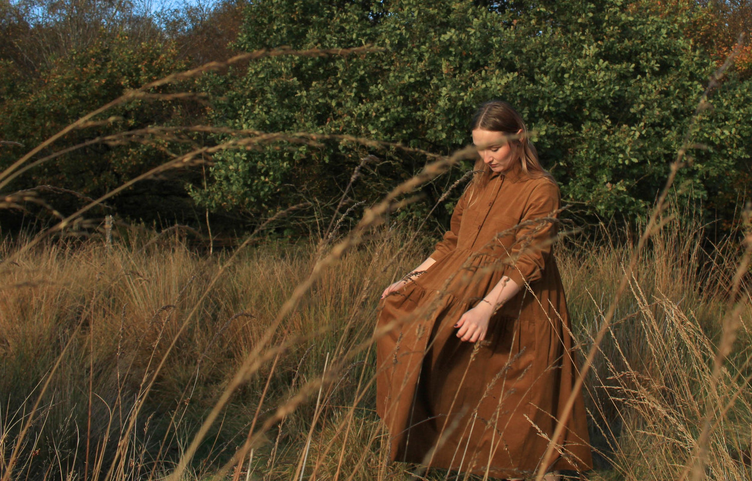 Woman in a brown dress standing in a field of tall grass with trees in the background
