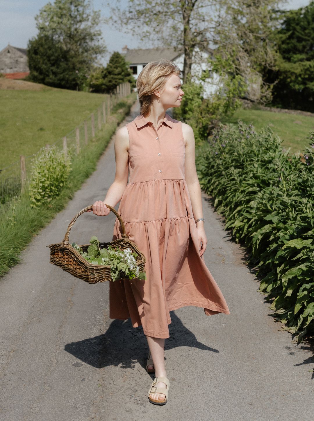 Woman in effie pink dress holding a basket of flowers walking on a path in a garden.