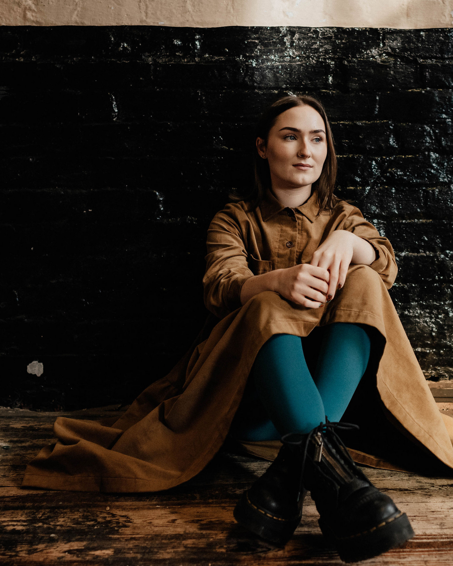 woman wearing a brown corduroy dress sat against a painted brick wall in old factory.