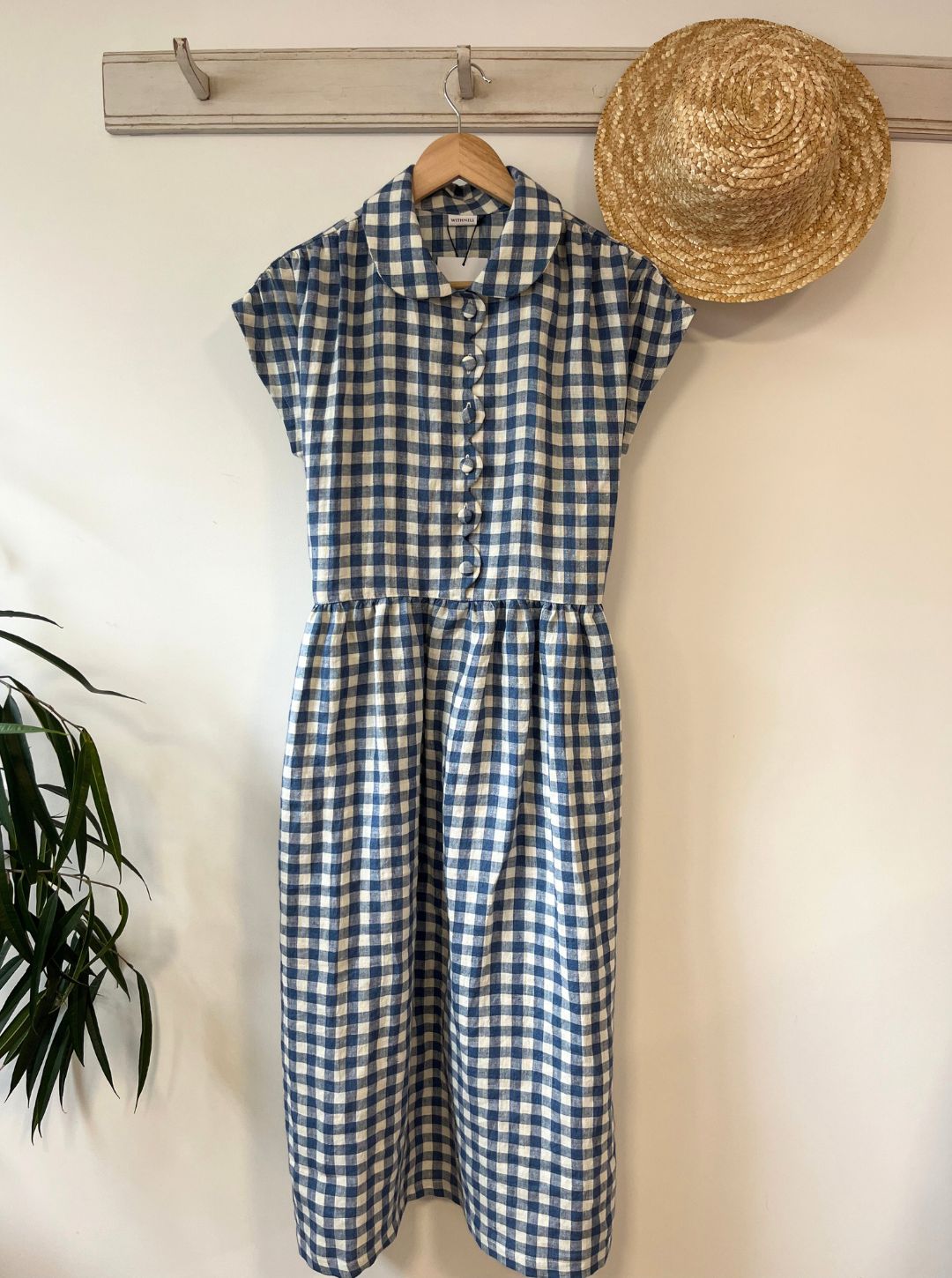 Blue and white checkered linen dress with a straw hat on a white wall.