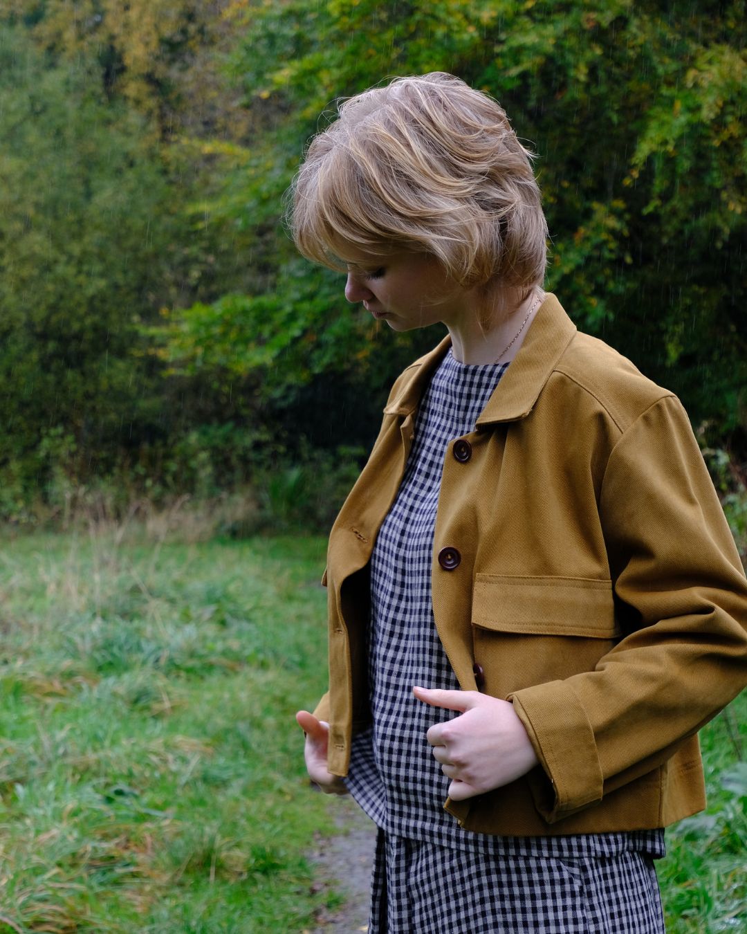Woman wearing a brown jacket in a natural setting with greenery.