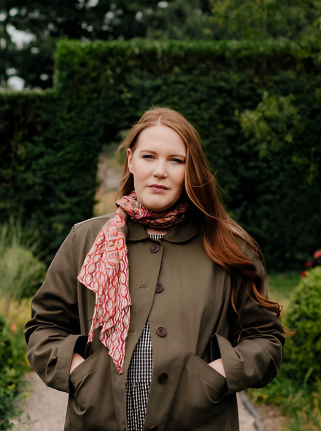 model wearing green hornbeam jacket and red patterned scarf standing in a garden.