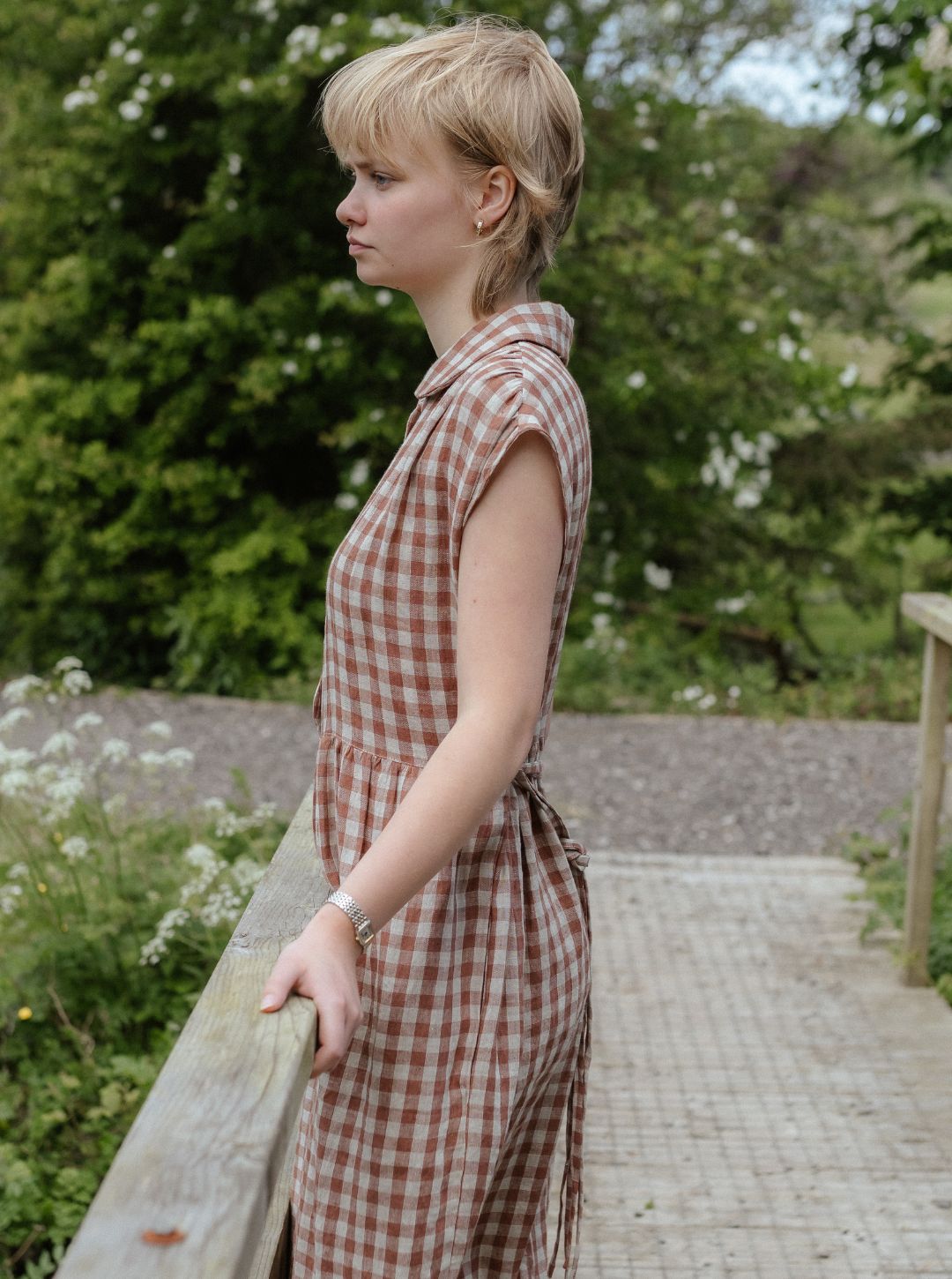 Woman in a checkered dress standing outdoors with greenery in the background