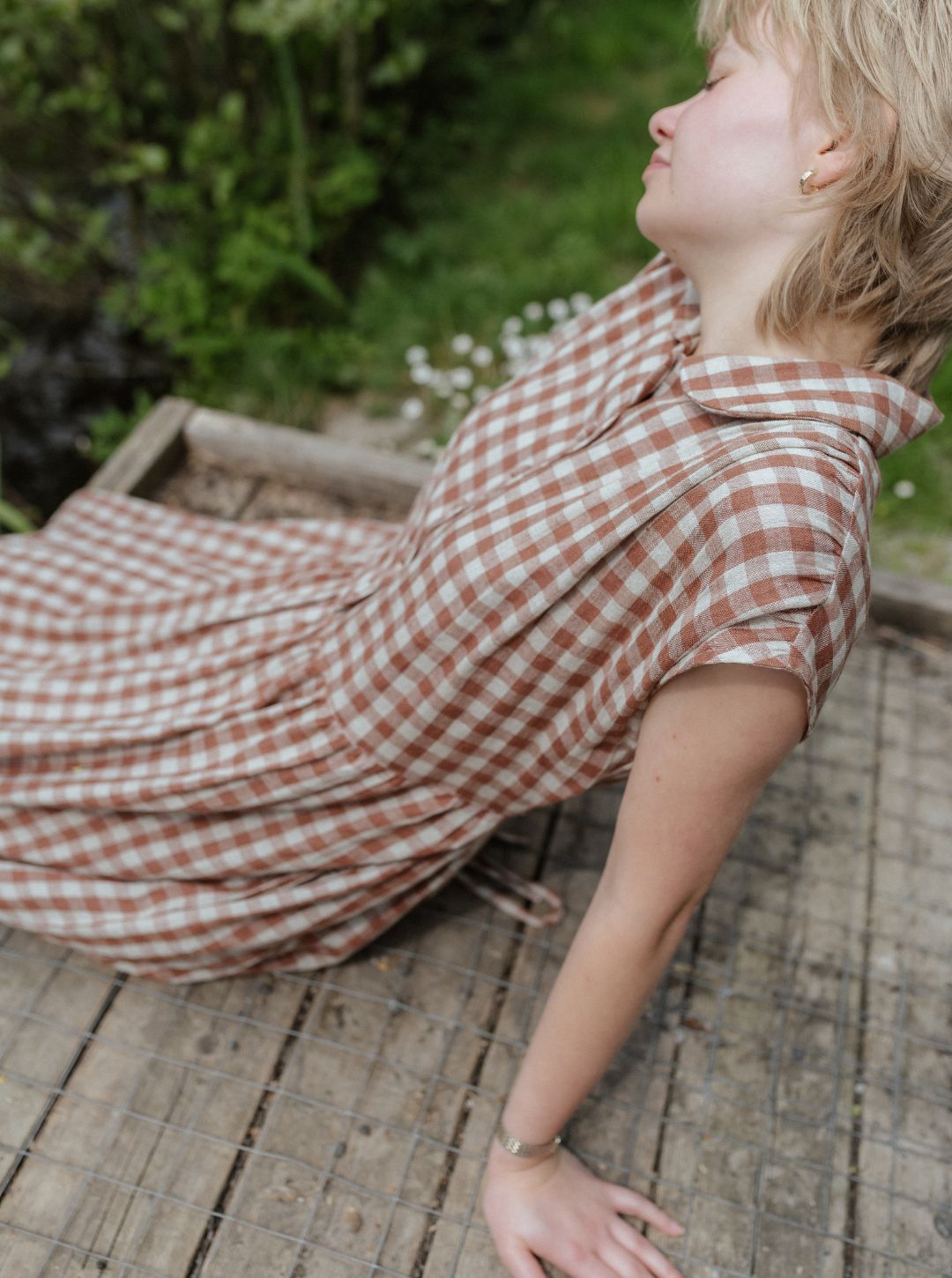 model in a brown and white checkered dress sitting on a wooden surface with greenery in the background