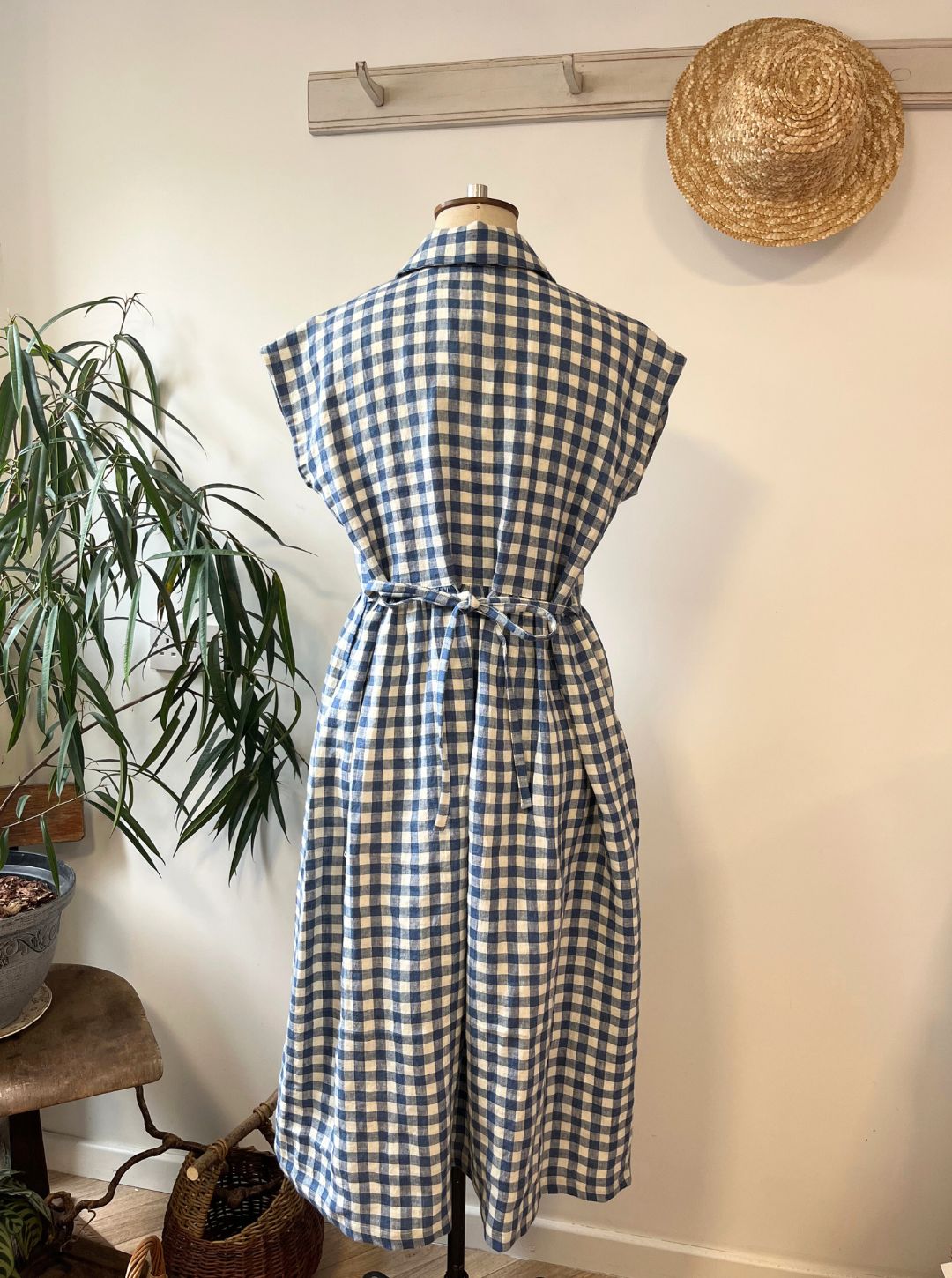 Blue and white checkered dress on a mannequin against a white wall with a plant and hat in the background.