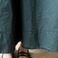 Close-up of a dark teal-green skirt  with visible fabric texture, hem stitching, and a small portion of a person's feet wearing black sandals on a wood floor.
