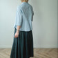 A woman viewed from behind wearing a light blue cropped blouse and a dark teal skirt, standing on a light wood herringbone floor against a plain off-white wall.