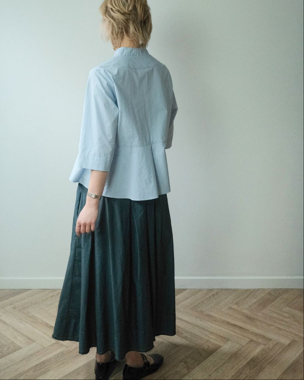 A woman viewed from behind wearing a light blue cropped blouse and a dark teal skirt, standing on a light wood herringbone floor against a plain off-white wall.