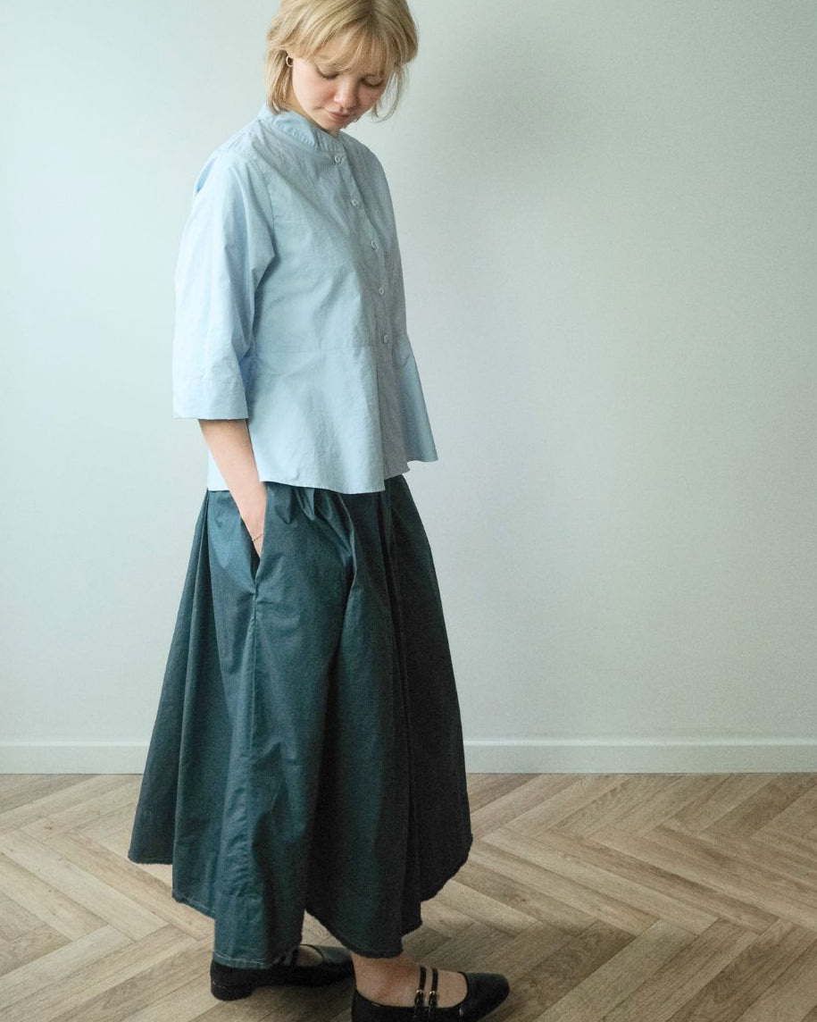 A woman stands in a minimalist room with a pale wall and light wood herringbone floor, wearing a light blue cropped button-up shirt, a dark teal-green skirt, and black Mary Jane flats.