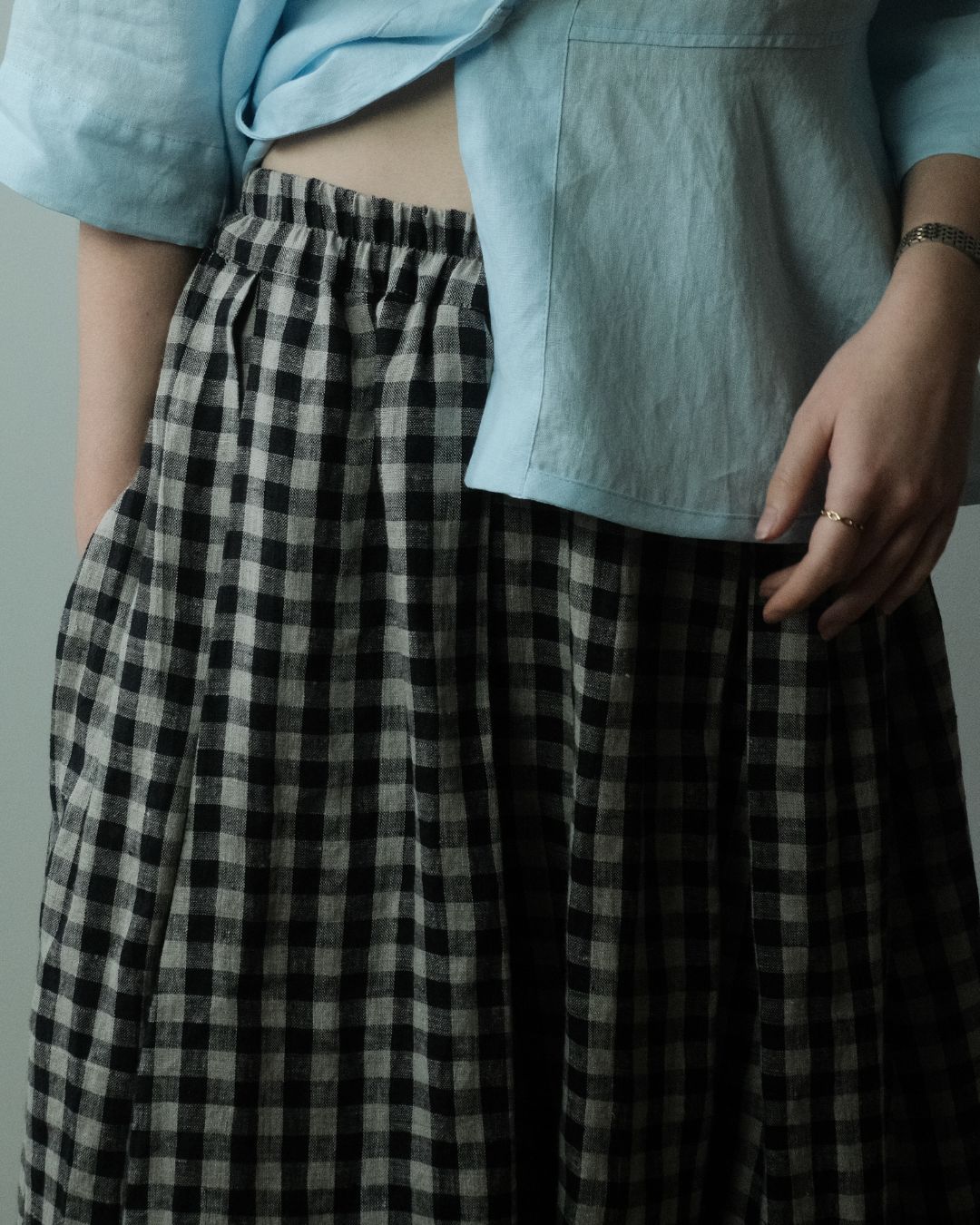 A close-up of a person wearing a black and white gingham check skirt with an elastic waistband, styled with a light blue cropped linen shirt.