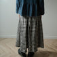A person standing indoors wearing a dark blue peplum top and a black-and-white gingham pleated skirt, paired with black shoes, against a plain light wall and wood floor.