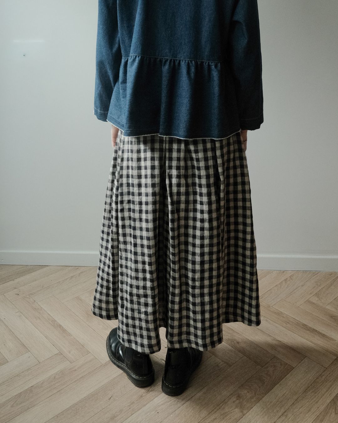 A person standing indoors wearing a dark blue peplum top and a black-and-white gingham pleated skirt, paired with black shoes, against a plain light wall and wood floor.