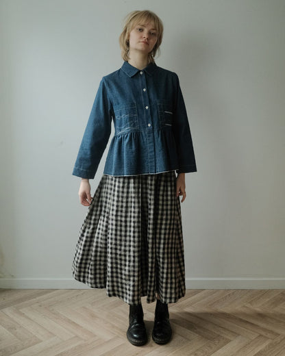A model stands in a minimalist room with a light gray wall and wood herringbone floor, wearing a dark blue denim long-sleeve button-up shirt tucked into a black and white gingham check midi skirt, paired with black leather boots.