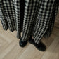 A person wearing a long black and beige gingham skirt and black leather slip-on shoes, standing on a light wood herringbone floor.