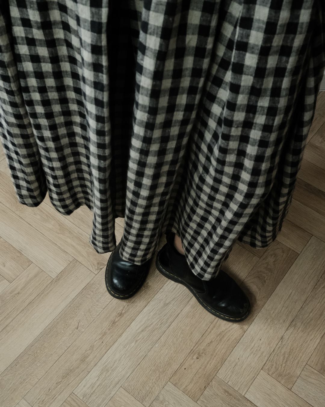 A person wearing a long black and beige gingham skirt and black leather slip-on shoes, standing on a light wood herringbone floor.