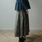 A person wearing a dark blue denim top and a black and white gingham pleated maxi skirt, paired with black leather ankle boots, standing on a light wood herringbone floor against a neutral gray wall.