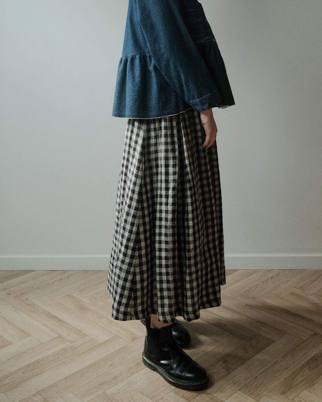 A person wearing a dark blue denim top and a black and white gingham pleated maxi skirt, paired with black leather ankle boots, standing on a light wood herringbone floor against a neutral gray wall.