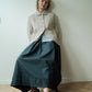 A woman leans against a plain off-white wall on a light wood herringbone floor, wearing a cream textured cardigan over a light blue collared shirt and a dark teal maxi skirt, paired with black Mary Jane flats.
