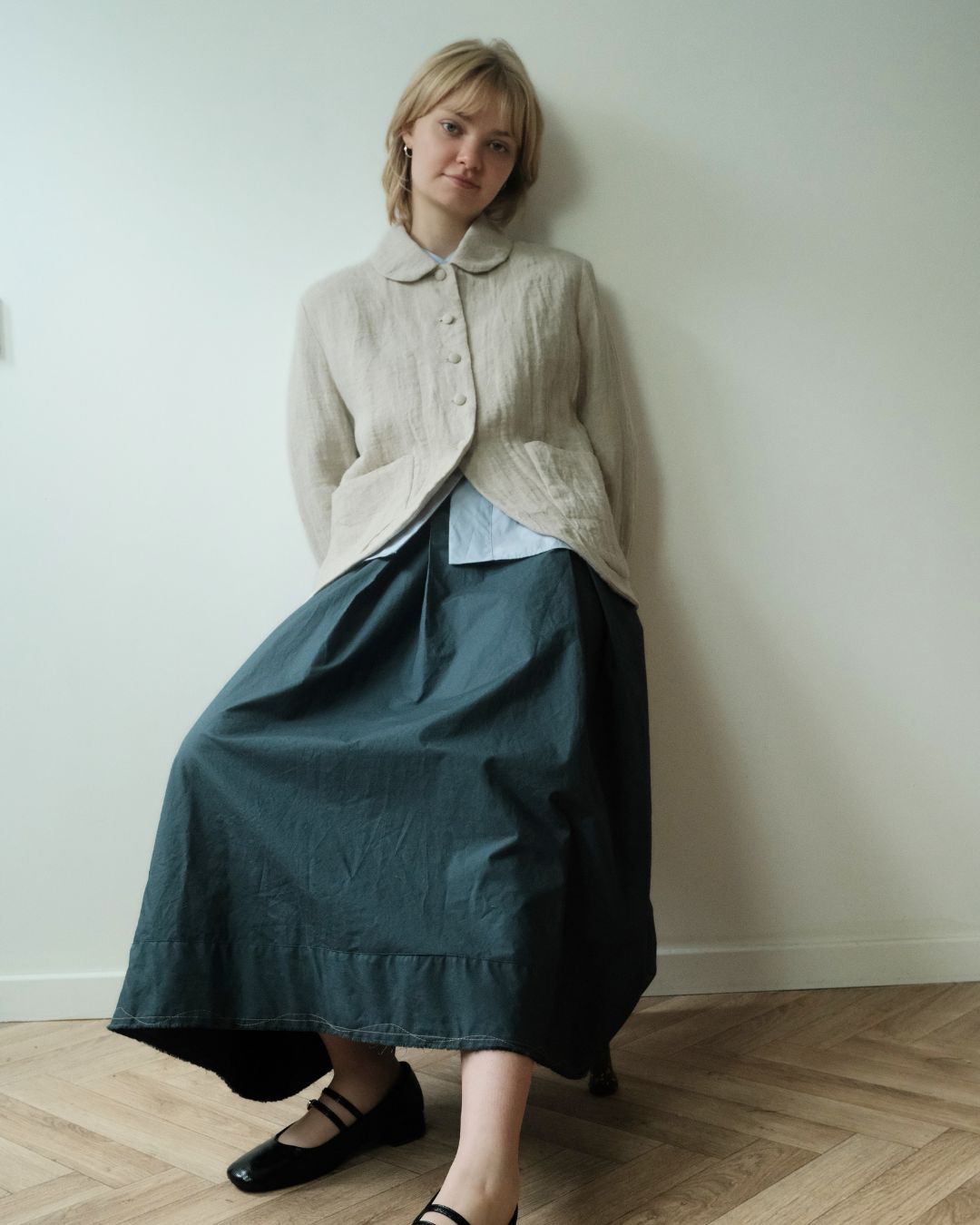 A woman leans against a plain off-white wall on a light wood herringbone floor, wearing a cream textured cardigan over a light blue collared shirt and a dark teal maxi skirt, paired with black Mary Jane flats.