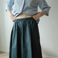 A person wearing a light blue button-up shirt and a dark teal-green gathered skirt, standing indoors against a plain light wall.