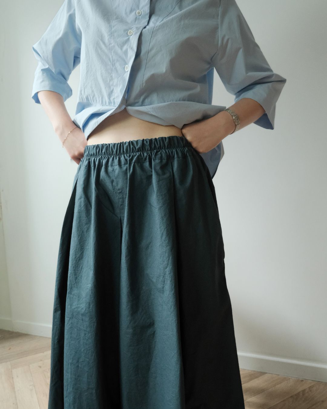 A person wearing a light blue button-up shirt and a dark teal-green gathered skirt, standing indoors against a plain light wall.