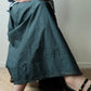 A person wearing a long, dark teal-gray linen midi skirt with visible contrast stitching, paired with a light blue shirt and black strappy heels, seated on a wooden floor against a plain wall.