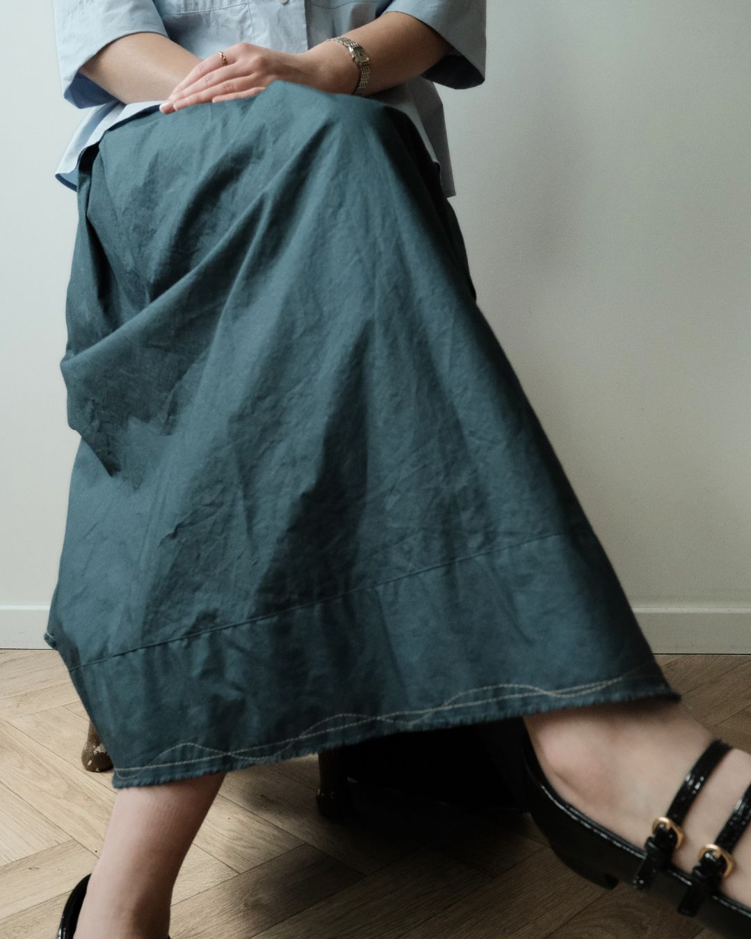 A person wearing a long, dark teal-gray linen midi skirt with visible contrast stitching, paired with a light blue shirt and black strappy heels, seated on a wooden floor against a plain wall.