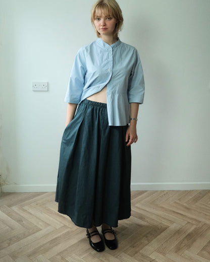 A woman stands in a room with a light gray wall and wood herringbone floor, wearing a light blue cropped button-up shirt and a dark teal wide-leg skirt.