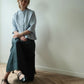 A woman sits on a wooden floor against a plain off-white wall, wearing a light blue short-sleeve button-up shirt and a dark green wide-leg skirt, paired with black Mary Jane shoes.