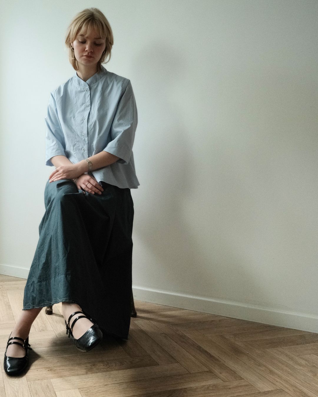 A woman sits on a wooden floor against a plain off-white wall, wearing a light blue short-sleeve button-up shirt and a dark green wide-leg skirt, paired with black Mary Jane shoes.