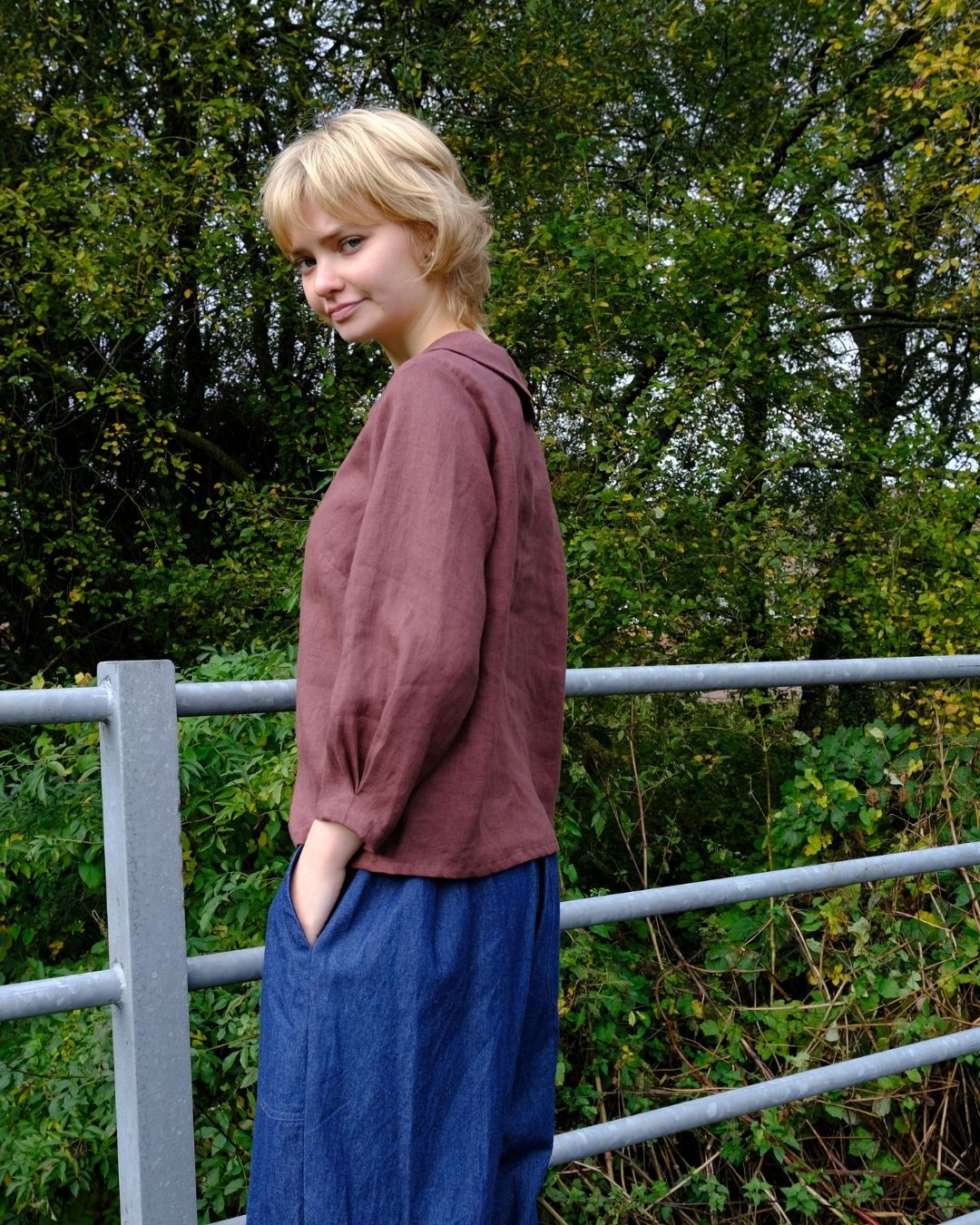 Woman wearing a maroon top and blue pants standing outdoors with greenery in the background