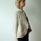 A woman in profile wearing a light beige linen jacket with a large front patch pocket, layered over a dark top and dark skirt, standing against a plain off-white wall.