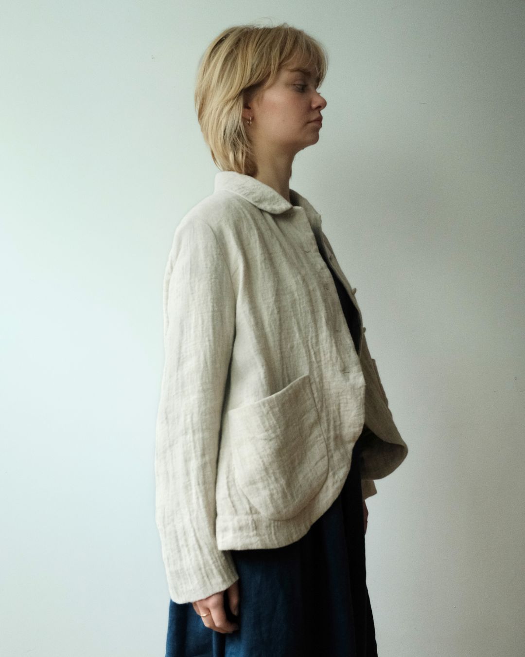 A woman in profile wearing a light beige linen jacket with a large front patch pocket, layered over a dark top and dark skirt, standing against a plain off-white wall.