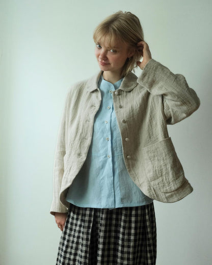 A woman wearing a light beige linen jacket over a light blue button-up shirt and a black-and-white gingham skirt, posing against a plain off-white wall.