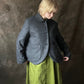 A person wearing a dark grey wool-jacket with a high collar and front buttons, paired with an olive green skirt, standing against a textured dark brown backdrop.