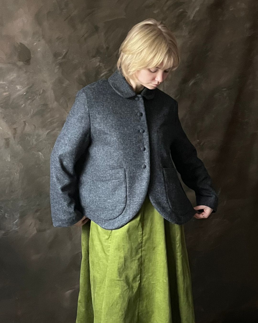 A person wearing a dark grey wool-jacket with a high collar and front buttons, paired with an olive green skirt, standing against a textured dark brown backdrop.