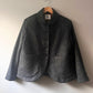 A dark gray textured wool jacket with a high folded collar and front buttons, hanging on a wooden hanger against a plain off-white wall.