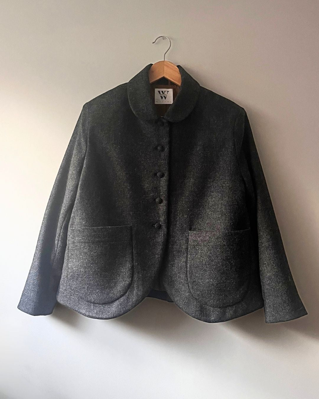 A dark gray textured wool jacket with a high folded collar and front buttons, hanging on a wooden hanger against a plain off-white wall.