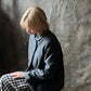 A woman with short blonde hair sits in profile wearing a dark charcoal gray button-up jacket and a black-and-white gingham skirt, posed against a textured gray backdrop.