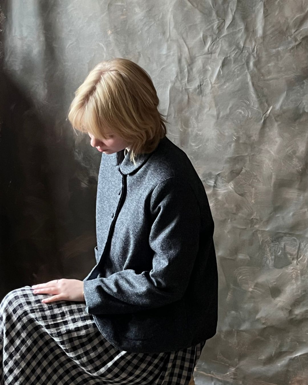 A woman with short blonde hair sits in profile wearing a dark charcoal gray button-up jacket and a black-and-white gingham skirt, posed against a textured gray backdrop.