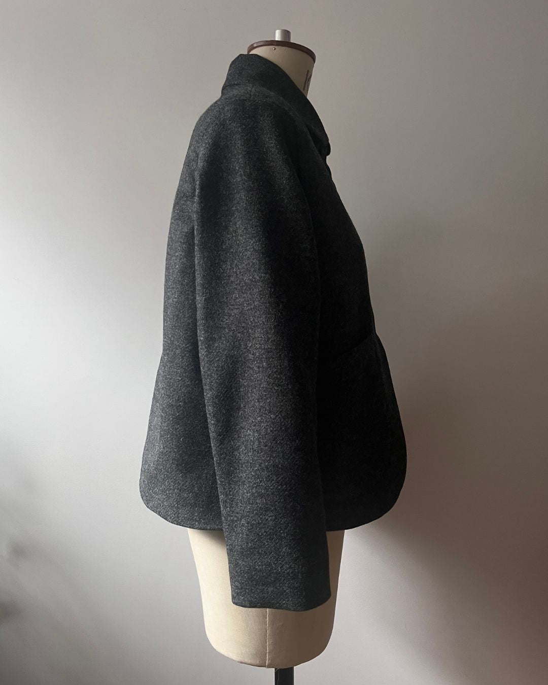 A dark gray textured wool jacket displayed on a cream-colored dress form mannequin against a neutral light gray wall.