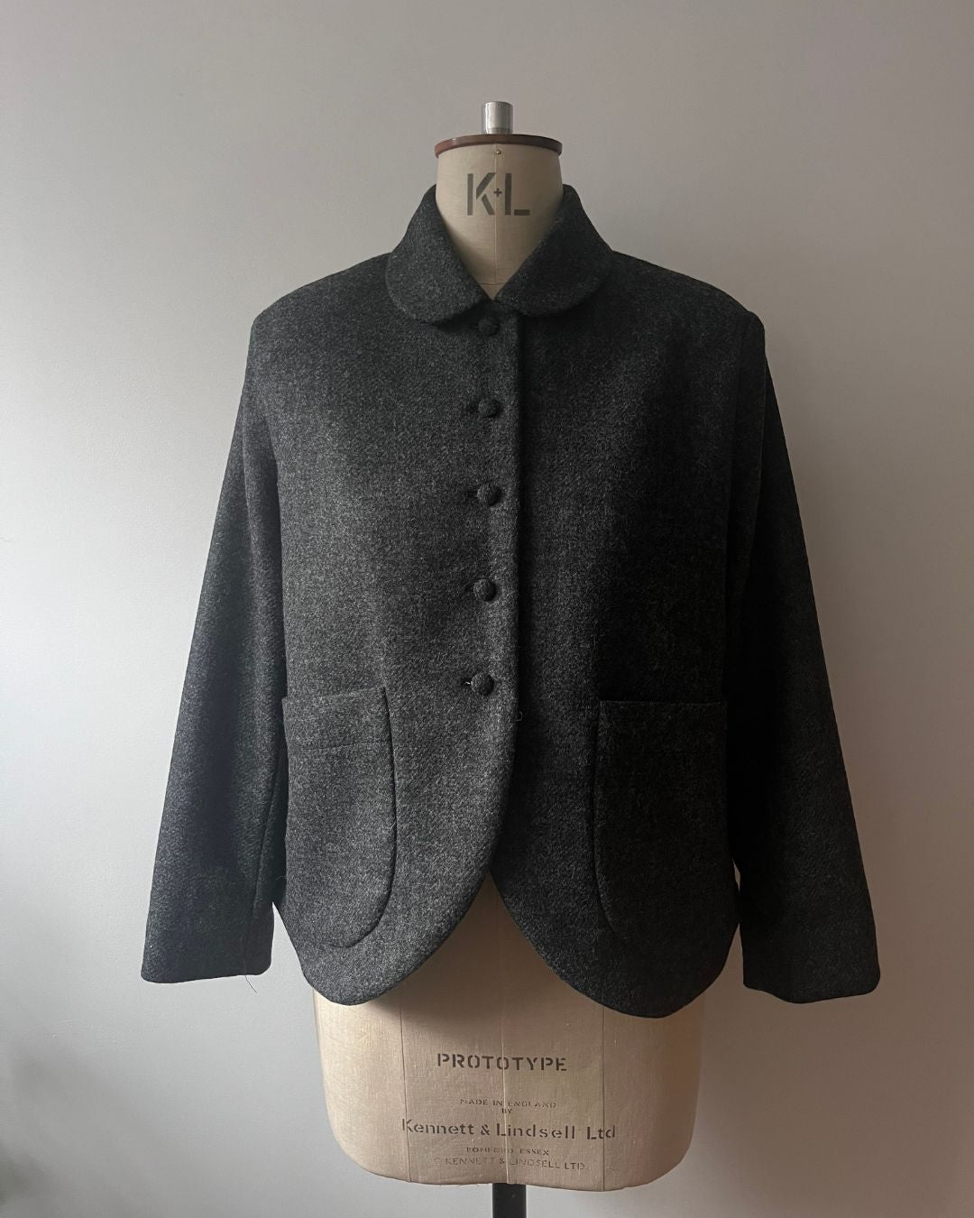 A dark gray textured wool jacket with a Peter Pan collar and front buttons, displayed on a vintage mannequin torso against a plain light gray wall.